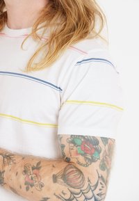 Valkoinen lyhythihainen t-paita, jossa on vaaleanpunaisia, sinisiä ja keltaisia vaakaraitoja. Paljaat käsivarret, joista näkyy värikkäitä tatuointeja kyynärvarressa.