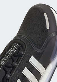 Närbild av en svart sneakers med vitt tre-stripes-logotyp, texturerat mesh-överdrag och svarta skosnören med tryckt text på tungan.