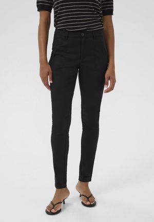 Femme portant un pantalon noir ajusté, un haut à manches courtes rayé et des sandales à talons noires, debout devant un fond blanc uni.