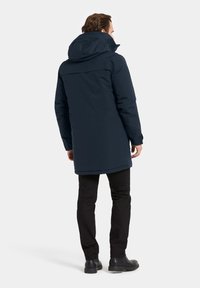 Marinblå vattentät parkas med huva, med en längre design, minimala sömmar och en slät textur. Bärs med svarta byxor och stövlar.