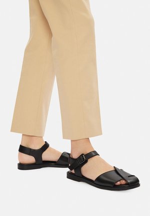 Černé kožené sandály s předním páskem, otevřenými špičkami a boční přezkou. Kombinované s béžovými zkrácenými kalhotami, představujícími minimalistický design.
