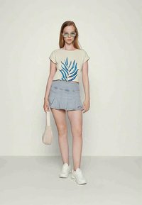 T-shirt bege com padrão de plantas, saia de denim plissada azul clara, sapatilhas brancas e uma pequena mala de ombro bege. Óculos verdes claros.