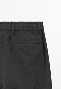 Détail d'un pantalon de costume gris foncé montrant la taille élastique à l'arrière, les passants de ceinture et une poche passepoilée unique sur le côté droit.