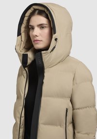 Beige Daunenjacke mit großer Kapuze, schwarzem Reißverschluss und Akzenten, gestepptem Design und zwei seitlichen Reißverschlusstaschen.
