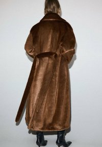 Cappotto lungo in pelo finto marrone con ampio colletto, vita cintata e tasche laterali. Il cappotto ha una texture morbida e una vestibilità ampia.