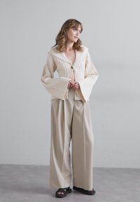 Cream ribbed jacket with wide sleeves and a front button, paired with loose beige trousers and black sandals. Simple, clean design.