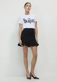 T-shirt bianco con la scritta "THE BEATLES" stampata in nero, abbinato a una gonna mini nera con texture, dotata di orlo arricciato e scarpe col tacco nere.