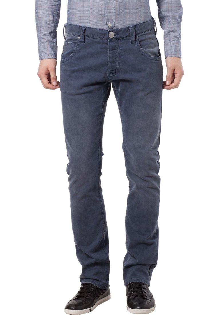 Mężczyzna w wyblakłych niebieskich dżinsach, czarnych trampkach i jasnoszarej koszuli w kratkę z długimi rękawami, stojący z relaksowanymi rękami.