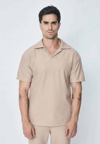 Chemise beige à manches courtes côtelées avec col plat et décolleté en V. Présente une coupe décontractée et un motif texturé subtil.