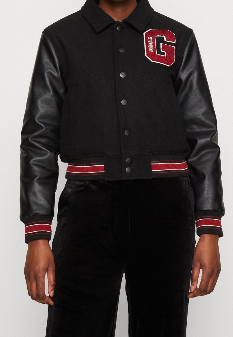 Svart kort jacka med yllekropp och läderärmar. Har en röd broderad "G"-patch och randiga ärmslut med röd och vit detaljering.