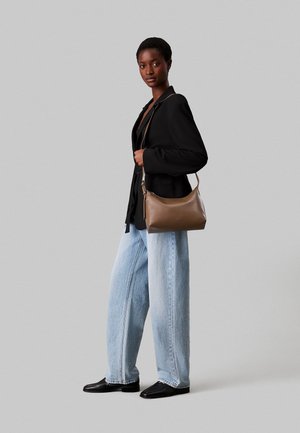 Borsa a spalla in pelle marrone con texture liscia e forma curvilinea, mostrata insieme a un modello in un blazer nero e jeans a gamba larga celesti.