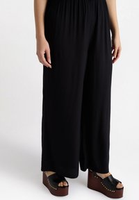 Svarta vida byxor med elastisk midja; kombinerade med kraftiga svarta platåsandaler med träsoffor och guldnitar.