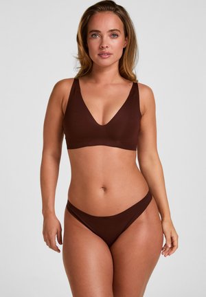 Frau mit heller Haut und langen blonden Haaren, die einen braunen BH mit V-Ausschnitt und passende Bikinihöschen trägt, stehend vor einem einfarbigen grauen Hintergrund.