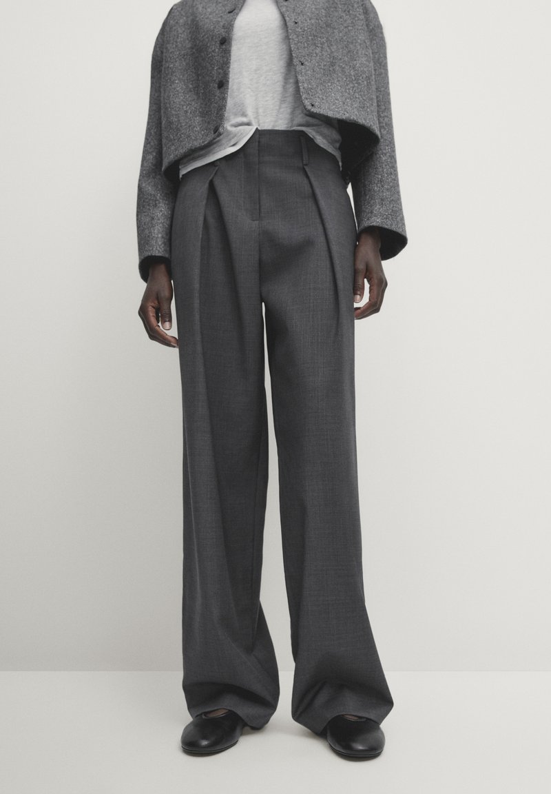 Persoon in donkere grijze hoge taille wijde broek, een lichtgrijs topje, een cropped grijs jack en zwarte platte schoenen tegen een effen achtergrond.