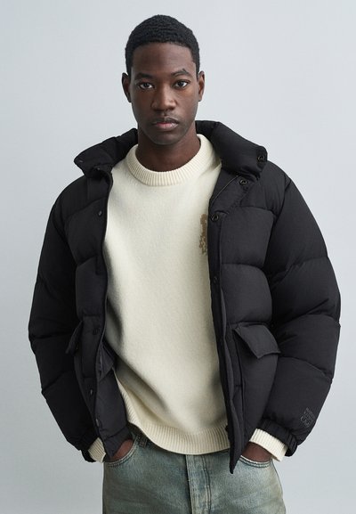 Mann, der eine schwarze Winterjacke über einem cremefarbenen Pullover und hellblauen Jeans trägt, steht mit den Händen in den Taschen vor einem schlichten Hintergrund.