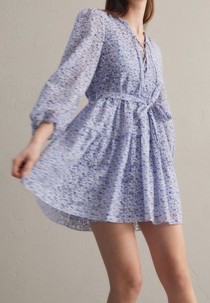 Woman wearing a light blue floral dress with puffed sleeves and a tie belt, captured mid-twirl against a plain background.