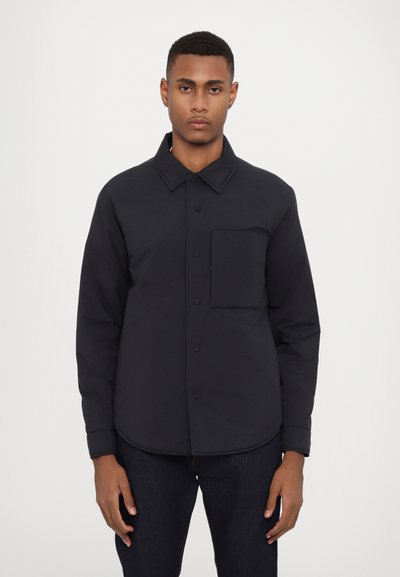Chaqueta negra de botonadura, hecha de tela suave, con cuello, bolsillo en el pecho y dobladillo redondeado. Combinada con pantalones vaqueros oscuros.