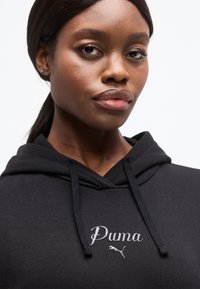 Mladá žena s tmavou pokožkou a vlasmi zviazanými vzadu, oblečená v čiernej mikine Puma s nastaviteľnými šnúrkami, na bielom jednoduchom pozadí.