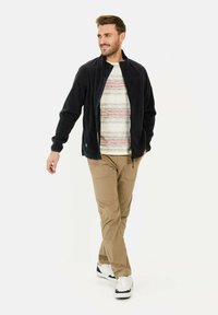 Zwarte fleece jas met hoge kraag, ritssluiting aan de voorkant en twee zijvakken. Wit gestreept shirt eronder, beige broek en witte sneakers.