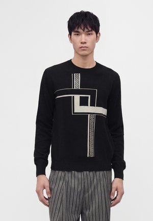 Ung mand iført en sort sweater med beige geometrisk mønster og grå bukser med tynde striber, stående foran en ensfarvet lys baggrund.