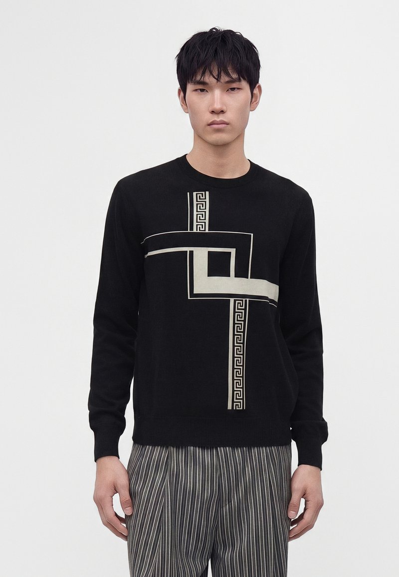 Jeune homme portant un pull noir avec un motif géométrique beige et un pantalon gris à fines rayures, debout devant un fond clair uni.