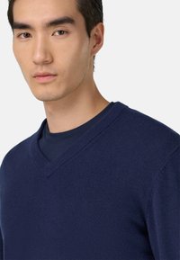 Pull bleu marine à col en V fabriqué en tissu doux, présentant une texture lisse et une coupe ajustée, porté sur une chemise foncée.