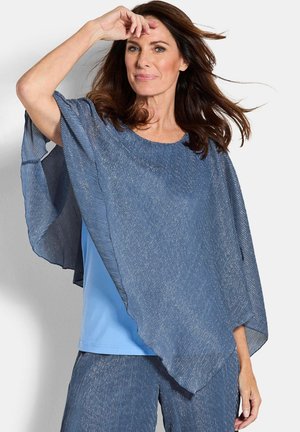 Frau trägt schimmerndes blaues Poncho und passende Hose, mit wehenden Haaren und erhobener Hand nahe der Stirn vor einfarbigem Hintergrund.