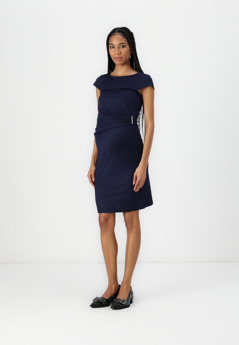 Vestido justo azul-marinho com mangas curtas, detalhe texturizado na cintura e acessórios em dourado. Combinado com sapatos pretos de bico fino.