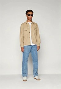 Camicia beige con bottoni sopra una maglietta bianca, jeans azzurri chiari e scarpe da ginnastica bianche. Il modello indossa occhiali da sole neri ed è in piedi contro uno sfondo neutro.