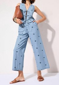 Žena ve světle modrém overalu bez rukávů s černými vyšívanými vzory, držící velkou hnědou pletenou tašku a obutá do hnědých sandálů.