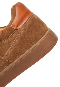 Hnědý semišový teniska s koženou patní záplatou v barvě světlého tan a silnou gumovou podrážkou, viditelné prošívání po stranách a vzadu.