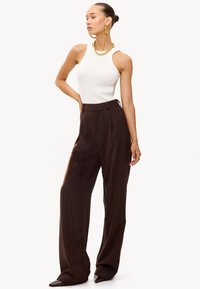 Top blanco sin mangas con textura acanalada combinado con pantalones de cintura alta marrones con rayas finas. Los accesorios destacados incluyen joyas doradas.