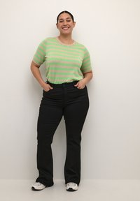 Pull à manches courtes rayé vert et beige, associé à un jean noir évasé et des baskets blanches, les mains sur les hanches, arrière-plan neutre.