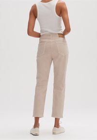 Pantalon en coton beige clair à coupe droite, avec poches arrière et une étiquette en cuir marron sur la ceinture, porté avec des chaussures blanches.