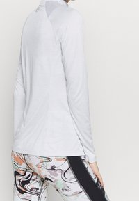 Weißes Langarmoberteil mit hohem Kragen, aus einem glatten, leichten Stoff. Dazu gemusterte Leggings mit bunten Wirbeln.