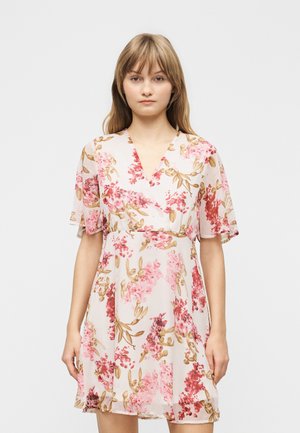 Giovane donna che indossa un vestito corto a maniche corte a fiori rosa e beige con scollo a V, in piedi davanti a uno sfondo chiaro e uniforme.