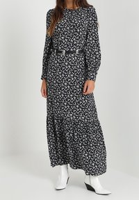 Květinové černé maxi šaty s dlouhými rukávy; mají fitted pas a vrstvenou sukni. Nosené s širokým černým opaskem a bílými kotníkovými botami.