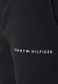 Černé bavlněné tepláky mají bílélogo "TOMMY" a červené a modré detaily na levé noze, jsou hladké na dotek a mají elastický pás.