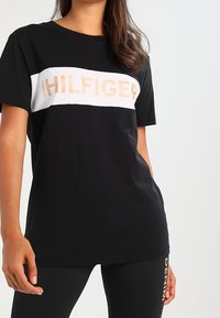 Černé bavlněné tričko s bílým a zlatým logem "TOMMY HILFIGER" na hrudi. Krátké rukávy a uvolněný střih.