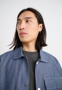 Denim jacket with a button-up front, light blue color, and a chest pocket, worn over a dark gray shirt, featuring a spread collar.