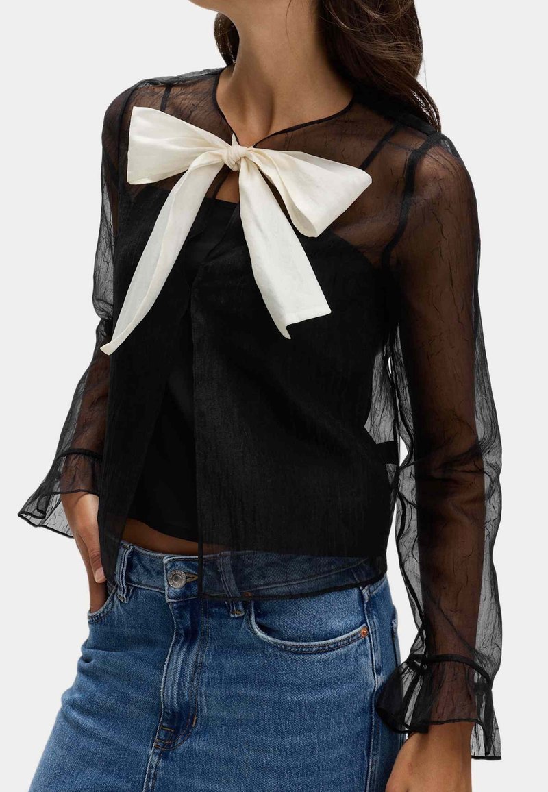 Femme portant un chemisier noir transparent à manches longues avec un grand nœud blanc au niveau du col, associé à un jean bleu.