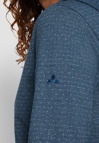 Sudadera con capucha de tejido azul oscuro con un sutil patrón moteado, que presenta un logotipo azul bordado en la manga izquierda. Material suave y texturado.
