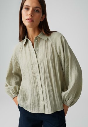 Jonge vrouw met steil bruin haar draagt een losse, lichtgroene blouse met textuur, knopen en opgeblazen mouwen, gecombineerd met een donkere broek, tegen een effen achtergrond.