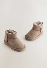 Tan mockasins av mockaläder med mjukt fuskpälsfoder, rund tå och strukturerad gummisula. Har en enkel rem med kardborre.