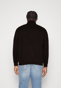 Veste noire à fermeture éclair avec un col montant, un tissu texturé et des poignets côtelés, associée à un jean en denim bleu. Vue arrière de l'ensemble.