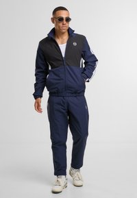 Námořnický sportovní komplet s černou horní částí bundy, zdobený bílými bočními pruhy a logem. Textilie vypadá hladce, má zapínání na zip.