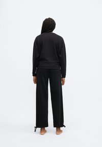 Črna jopa in ujemajoče široke hlače. Gladek material, sproščen kroj in zavezovanje na hlačnicah. Pogled od zadaj.