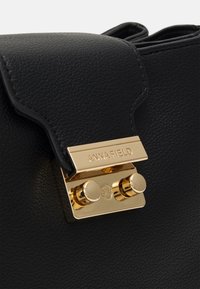 Černá kabelka z texturované kůže s zapínáním v zlaté barvě, zdobeným vygravírovaným logem a kulatými kovovými doplňky.