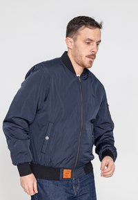 Marine bomberjack met ribgebreide manchetten en zoom, ritssluiting aan de voorkant, zijzakken en een oranje label op de taille. Soepele stoftextuur.