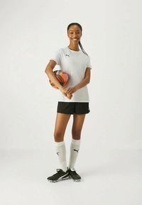 Camisa blanca de Puma, pantalones cortos negros, calcetas blancas y botines negros. Sosteniendo un balón de fútbol naranja y negro. Posando sonriente, fondo neutral.